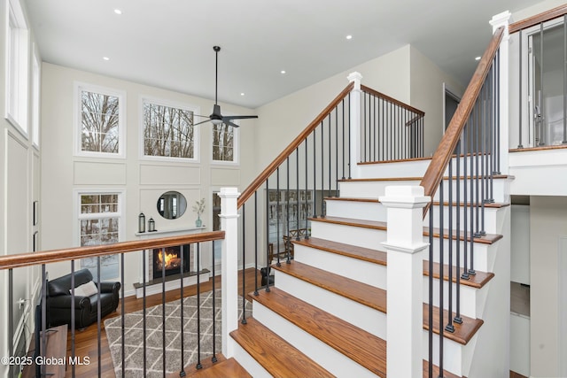 staircase with wood finished floors, ceiling fan, a glass covered fireplace, a towering ceiling, and recessed lighting