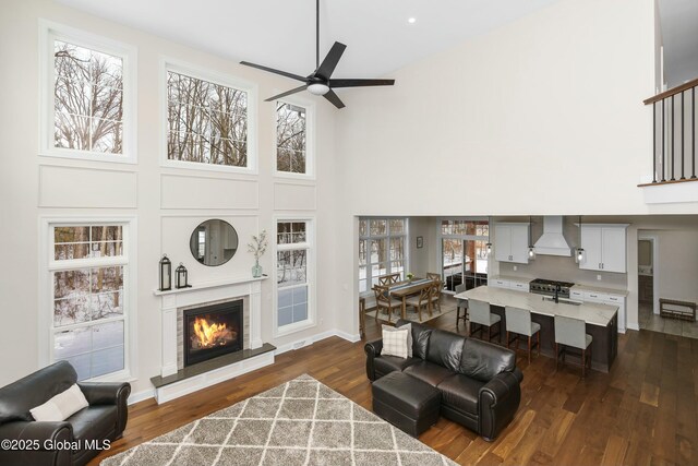 living area with a high ceiling, ceiling fan, dark wood-style floors, a glass covered fireplace, and baseboards