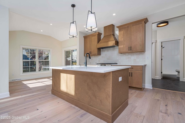 kitchen with premium range hood, a center island with sink, brown cabinetry, light wood-type flooring, and lofted ceiling