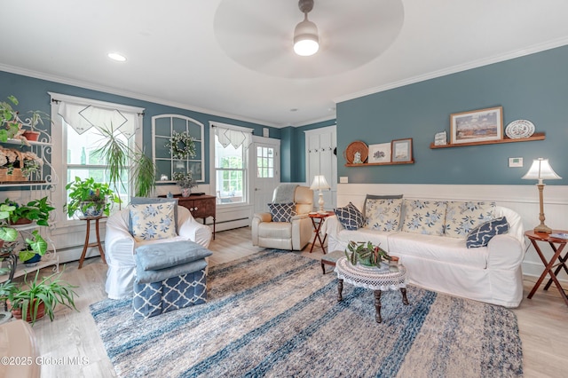 living area featuring baseboard heating, crown molding, wood finished floors, ceiling fan, and wainscoting