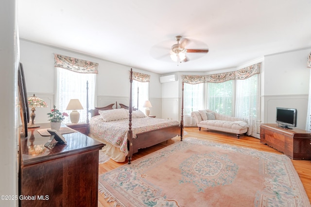 bedroom featuring multiple windows, a wainscoted wall, light wood-type flooring, a wall unit AC, and a ceiling fan