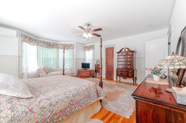 bedroom featuring light wood-type flooring, a decorative wall, wainscoting, an AC wall unit, and a ceiling fan