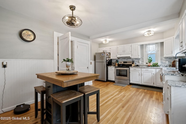 kitchen with appliances with stainless steel finishes, light wood finished floors, a wainscoted wall, exhaust hood, and white cabinetry