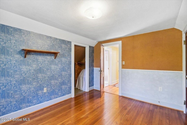 empty room featuring lofted ceiling, hardwood / wood-style floors, a textured ceiling, baseboards, and wallpapered walls