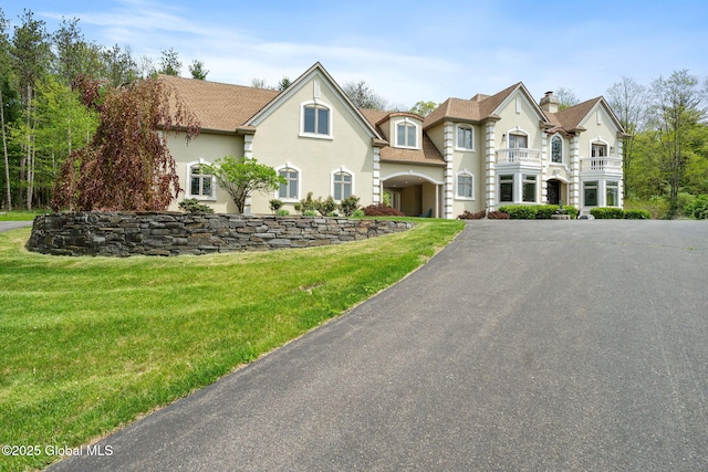 view of front facade with a balcony, stucco siding, driveway, a front yard, and a chimney