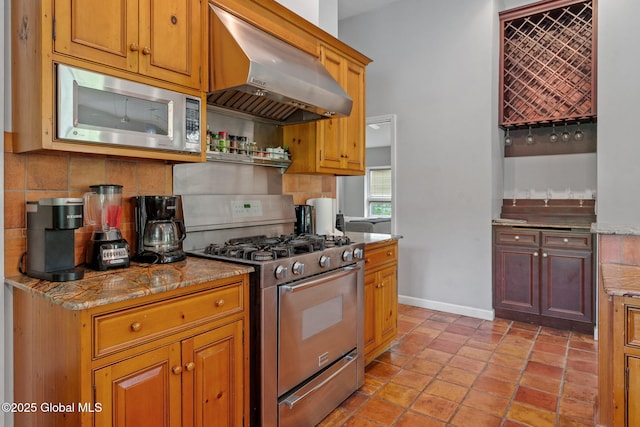 kitchen featuring appliances with stainless steel finishes, under cabinet range hood, decorative backsplash, baseboards, and brown cabinets