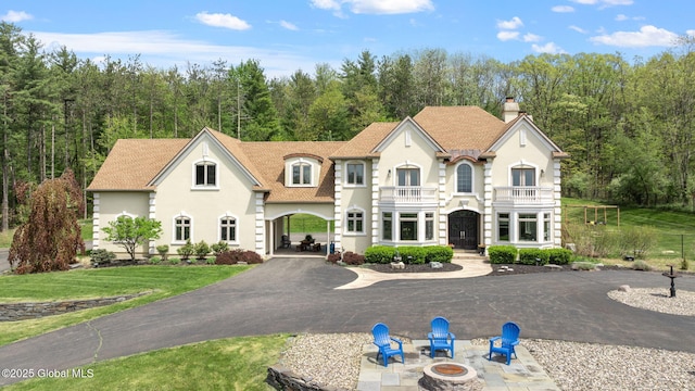 french country inspired facade featuring an outdoor fire pit, stucco siding, a chimney, a balcony, and a wooded view
