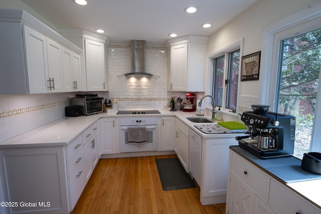 kitchen with white oven, wall chimney range hood, black electric stovetop, a sink, and white cabinets