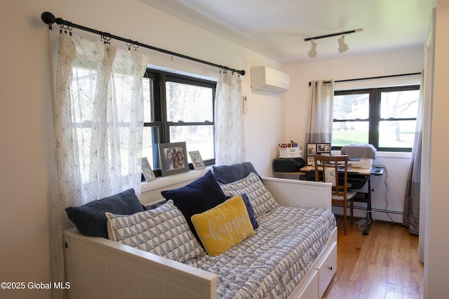 bedroom with baseboard heating, a wall mounted air conditioner, wood finished floors, and multiple windows