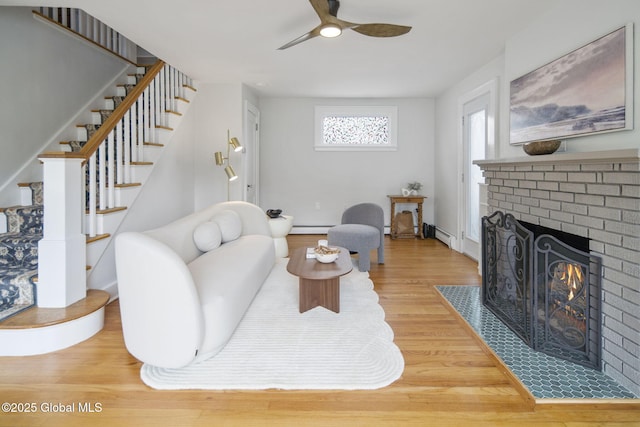 living room with a ceiling fan, a fireplace, wood finished floors, a baseboard heating unit, and stairway