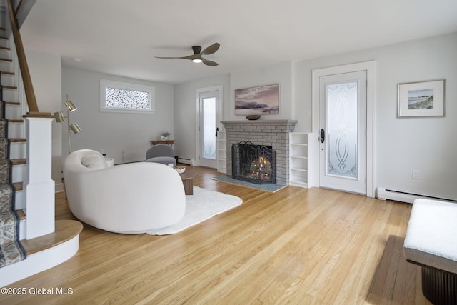 living area featuring baseboard heating, a ceiling fan, stairway, a brick fireplace, and wood finished floors