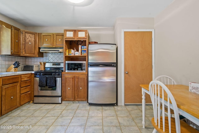 1927 Berne Altamont Rd, Altamont, NY 12009 - kitchen featuring stainless steel appliances, under cabinet range hood, tile countertops, decorative backsplash, and brown cabinetry
