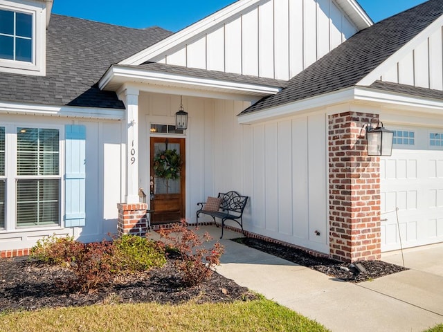 doorway to property with an attached garage, brick siding, board and batten siding, and roof with shingles