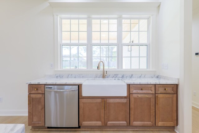 kitchen with light wood-type flooring, light stone countertops, brown cabinetry, stainless steel dishwasher, and a sink