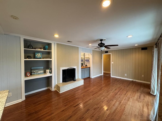 unfurnished living room featuring crown molding, ceiling fan, built in features, a fireplace, and dark hardwood / wood-style flooring