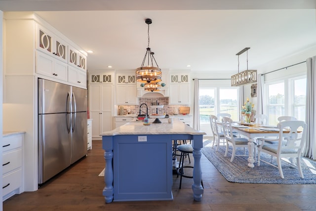 kitchen with white cabinetry, tasteful backsplash, stainless steel fridge, decorative light fixtures, and a center island with sink