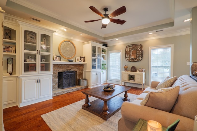 living room featuring ceiling fan, dark wood-type flooring, a raised ceiling, a stone fireplace, and crown molding