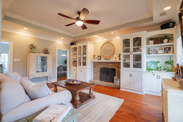 living room featuring a tray ceiling, ceiling fan, crown molding, hardwood / wood-style floors, and a stone fireplace