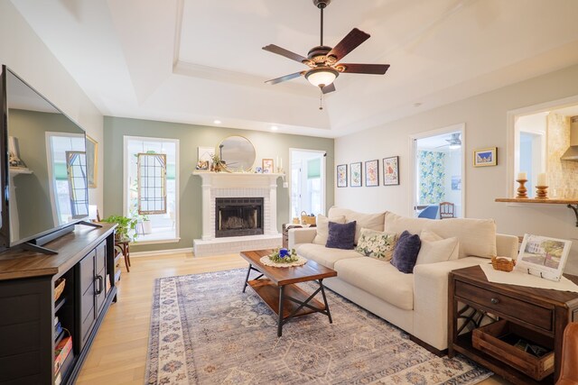 living room with a brick fireplace, ceiling fan, baseboards, light wood-type flooring, and a tray ceiling