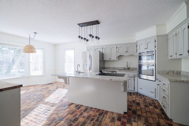 kitchen featuring appliances with stainless steel finishes, an island with sink, brick floor, and pendant lighting