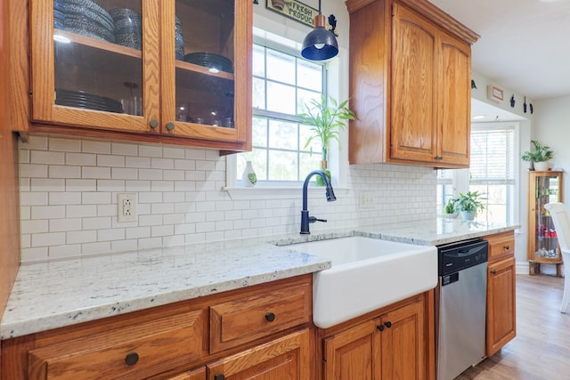 kitchen featuring glass insert cabinets, a sink, brown cabinets, light stone countertops, and stainless steel dishwasher