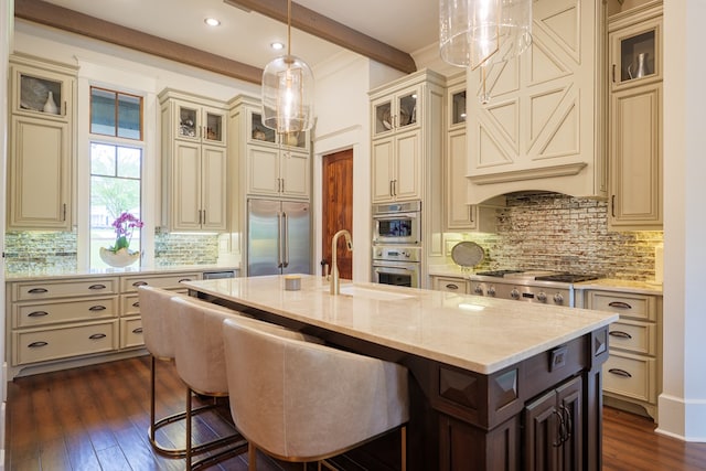 kitchen with stainless steel appliances, dark wood-type flooring, cream cabinets, a center island with sink, and beamed ceiling