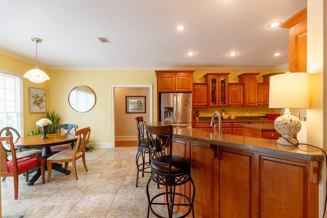 kitchen with a kitchen bar, stainless steel fridge, ornamental molding, dark countertops, and brown cabinets