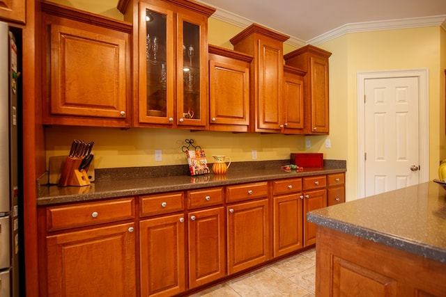 kitchen featuring crown molding, dark countertops, glass insert cabinets, brown cabinetry, and light tile patterned floors