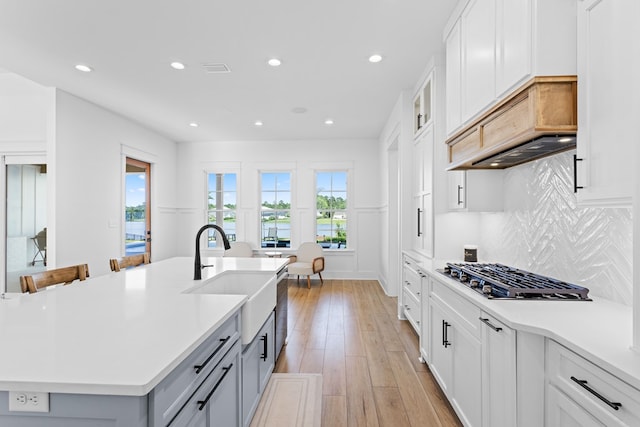 kitchen featuring stainless steel gas stovetop, a kitchen island with sink, white cabinets, sink, and light hardwood / wood-style flooring