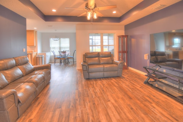 living area with ceiling fan, a tray ceiling, baseboards, and wood finished floors