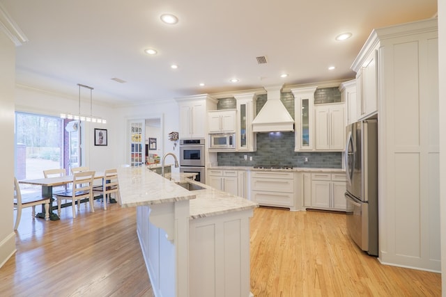 kitchen featuring premium range hood, a kitchen island with sink, hanging light fixtures, stainless steel appliances, and white cabinets