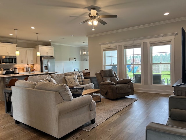 living room featuring wood-type flooring, ceiling fan with notable chandelier, crown molding, and sink