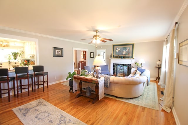 living room featuring light wood finished floors, a glass covered fireplace, ceiling fan with notable chandelier, baseboards, and ornamental molding
