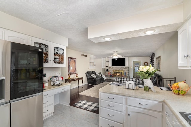 kitchen with stainless steel fridge with ice dispenser, a stone fireplace, a peninsula, ceiling fan, and light wood-style flooring