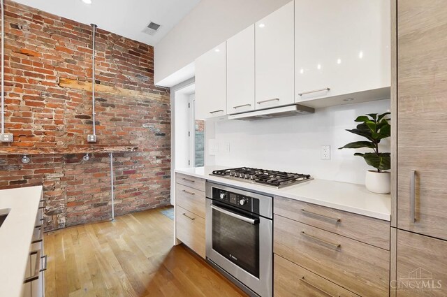 kitchen featuring brick wall, under cabinet range hood, light wood-style floors, visible vents, and appliances with stainless steel finishes