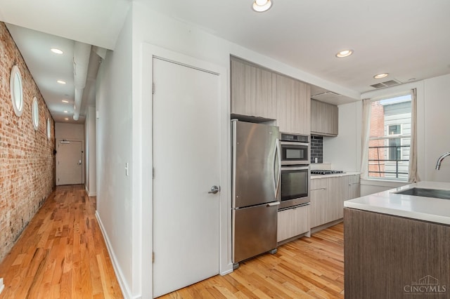 kitchen with appliances with stainless steel finishes, a sink, brick wall, light wood-style floors, and light countertops