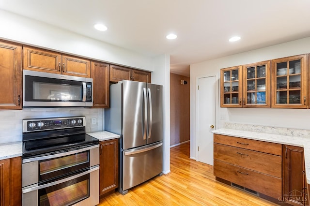 kitchen featuring appliances with stainless steel finishes, backsplash, glass insert cabinets, light wood-type flooring, and light stone countertops