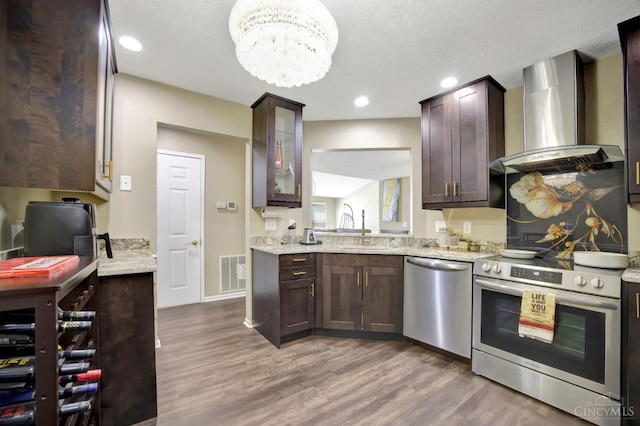 kitchen featuring appliances with stainless steel finishes, wall chimney exhaust hood, dark wood-type flooring, dark brown cabinetry, and a chandelier