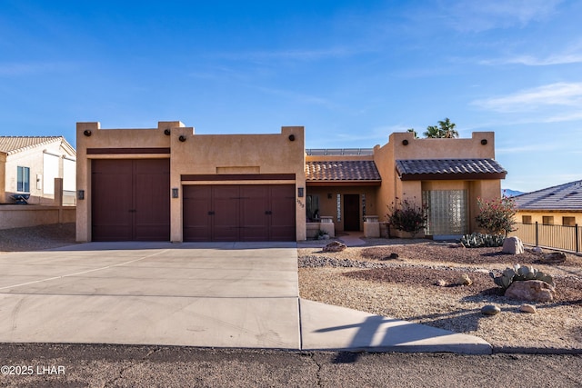 pueblo-style house featuring a garage, a tile roof, fence, driveway, and stucco siding