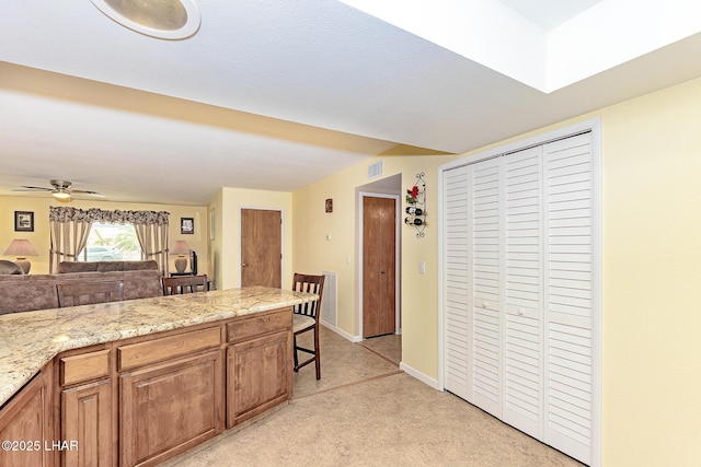 kitchen featuring a ceiling fan, visible vents, baseboards, light stone countertops, and brown cabinetry