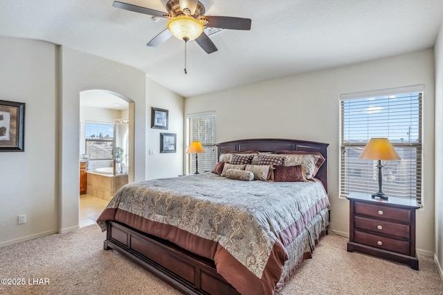 bedroom featuring light carpet, baseboards, arched walkways, a ceiling fan, and ensuite bathroom