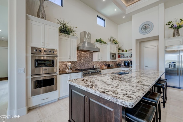 kitchen with tasteful backsplash, a towering ceiling, built in appliances, wall chimney range hood, and a sink