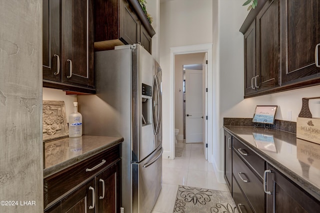 kitchen with light tile patterned floors, baseboards, dark brown cabinets, stainless steel fridge with ice dispenser, and dark stone countertops