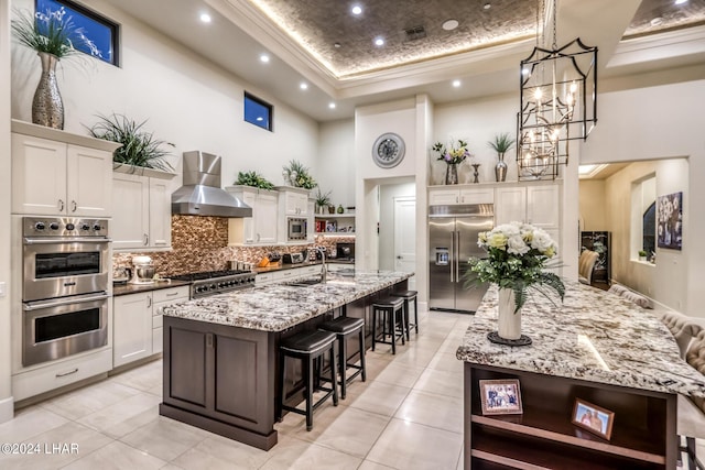 kitchen featuring a raised ceiling, a large island, built in appliances, wall chimney range hood, and open shelves