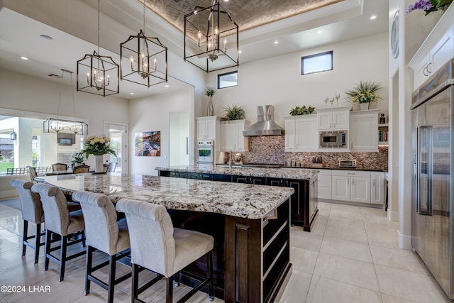 kitchen with a tray ceiling, open shelves, wall chimney range hood, built in appliances, and a large island with sink