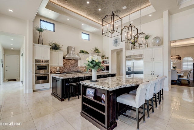 kitchen featuring a tray ceiling, open shelves, appliances with stainless steel finishes, a large island with sink, and wall chimney exhaust hood