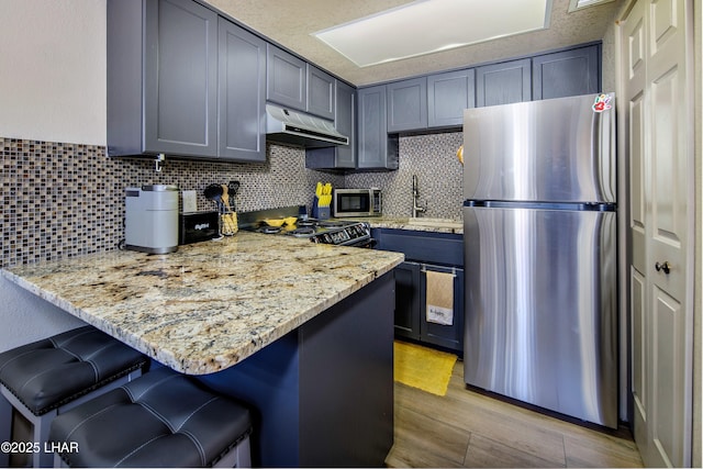kitchen with a sink, under cabinet range hood, a kitchen breakfast bar, light stone counters, and appliances with stainless steel finishes