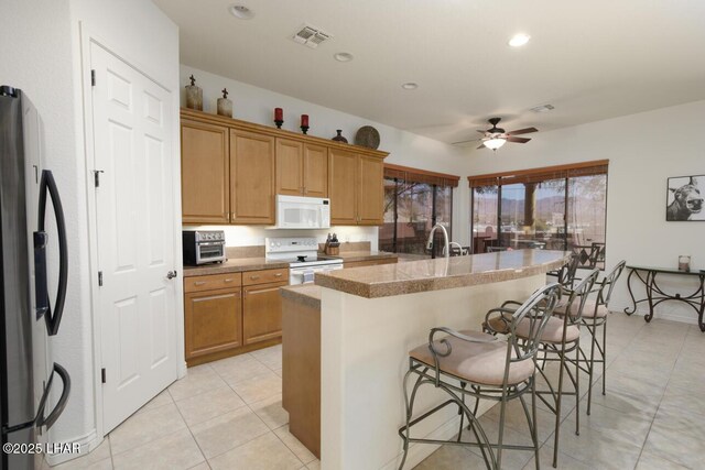kitchen with white appliances, a center island with sink, visible vents, a breakfast bar area, and brown cabinets