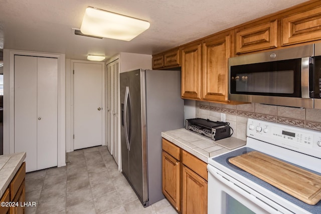 kitchen with light tile patterned floors, brown cabinetry, appliances with stainless steel finishes, tile counters, and backsplash