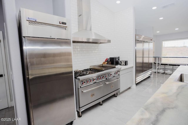 kitchen with high end appliances, recessed lighting, white cabinetry, wall chimney range hood, and tasteful backsplash